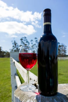 A red wine bottle and glass resting on a white fence in the country.