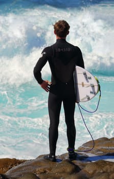 CAPE SOLANDER, AUSTRALIA - AUGUST -, 2015; Australian surfer stands and watches the wavesat Cape Solander.  He is wearing a RipCurl wetsuit and carrying his surfboard