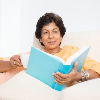 Portrait of a 50s Indian mature woman reading book and drinking coffee at home. Indoor senior people living lifestyle.