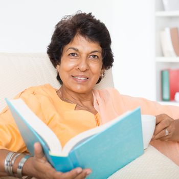 Portrait of a 50s Indian mature woman reading book and drinking coffee at home. Indoor senior people living lifestyle.