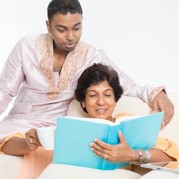 Portrait of happy Indian family reading book, sharing hobby at home. Beautiful mature 50s Indian mother and her 30s grown son.