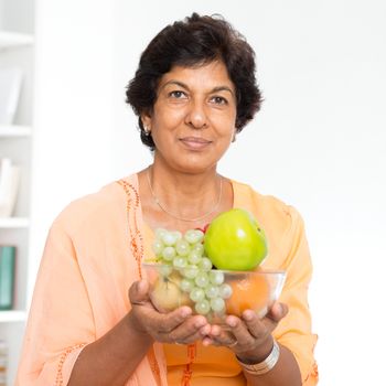 Old people healthy eating. Portrait of a 50s Indian mature woman holding fresh fruits at home. Indoor senior people living lifestyle.