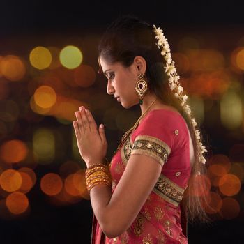 Indian woman in traditional sari praying and celebrating Diwali or deepavali, fesitval of lights at temple. Female prayer hands folded, beautiful lights bokeh background.