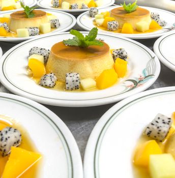 Many caramel, custard, vanilla flavored pudding on white plate with diced dragon fruit, diced peach, cantaloupe and a mint leaf