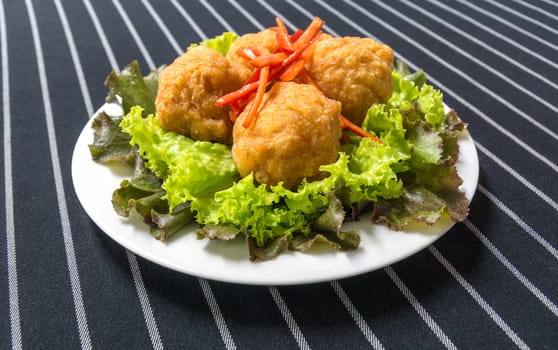 Deep fried shrimp balls, garnished with sliced red chili, red oak and green lettuce on a white plate on dark blue background with white stripe. ingredients-shrimp meat, pork fat, pepper, salt, sugar