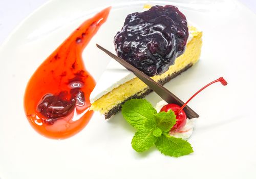 Cheese cake dressing with white cream, blue berry jam, red berry jam, red cherry, mint leaves and chocolate on a plate on white background