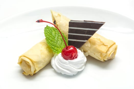 apple strudel decorating with topping cream, a red cherry, a fresh green mint leaf, chocolate on white plate on white background