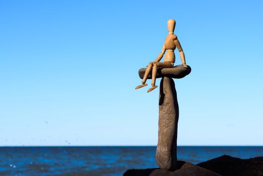 Wooden mannequin sitting on the top of sea stone