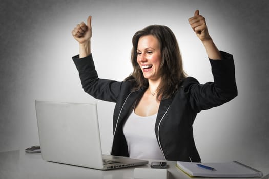 Happy woman sitting in office at desk in front of her laptop and holding her thumbs up