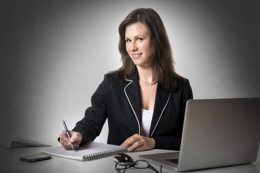 Smiling Businesswoman sitting at desk in office, writing something on a pad, isolated on white background