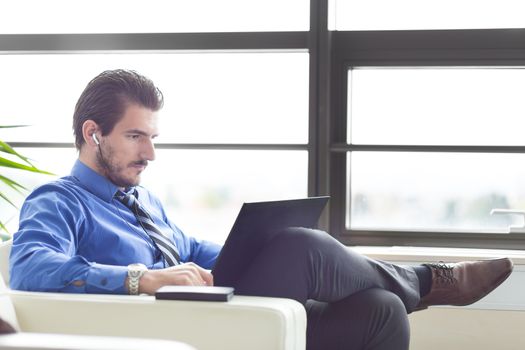 Busy, focused businessman in office working on his laptop wearing headphones. Side view. 