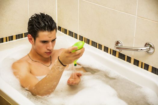 Handsome young man in bathtub at home having bath, washing body and hair with bathfoam and shampoo