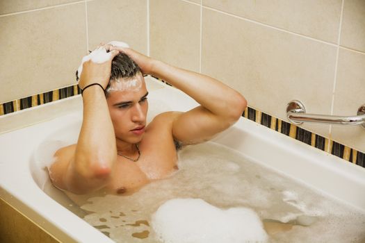 Handsome young man in bathtub at home having bath, washing body and hair with bathfoam and shampoo