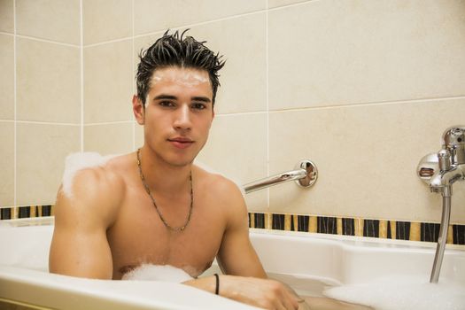 Handsome young man in bathtub at home having bath, washing body and hair with bathfoam and shampoo