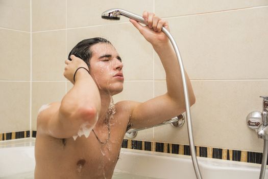 Handsome young man in bathtub at home having bath, washing body and hair with bathfoam and shampoo