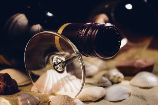 An unopened bottle of red wine resting on a glass on sand and shells. Film look, grain effect.