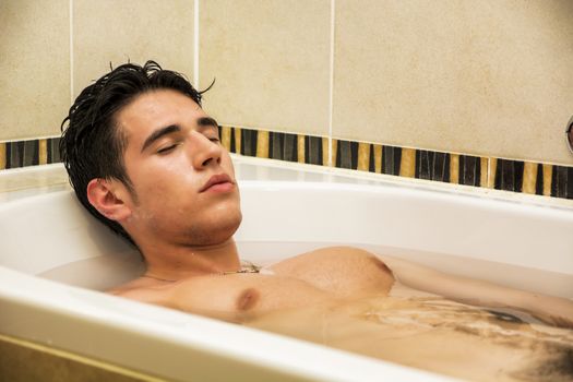 Handsome young man in bathtub at home having bath, washing body and hair with bathfoam and shampoo