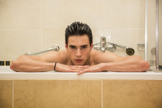 Handsome young man in bathtub at home having bath, leaning on tub edge, looking at camera