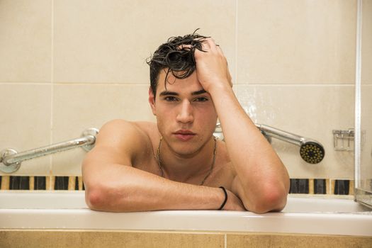 Handsome young man in bathtub at home having bath, leaning on tub edge, looking at camera