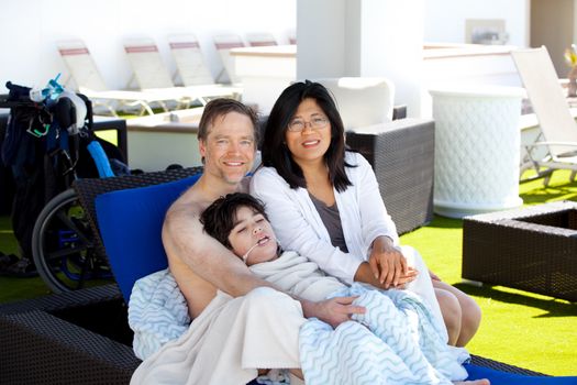 Multiracial couple with young disabled son on blue poolside lounger, drying off after swimming. Child has cerebral palsy. 