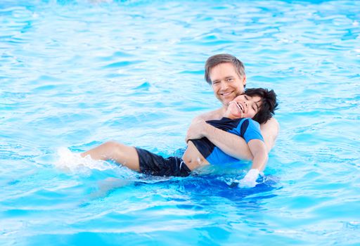 Caucasian father swimming in pool with biracial disabled son in his arms. Child has cerebral palsy.