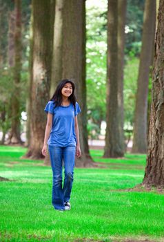 Young teen biracial Asian  girl walking under tall trees, on bright green grass