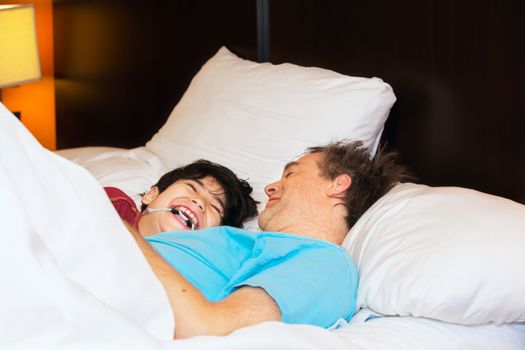 Caucasian father taking a nap with young disabled biracial son, in bed, laughing and talking together. Child has cerebral palsy. 