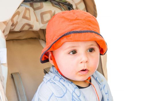 Baby with orange cap sitting in a stroller, looking amazed. Background isolated.