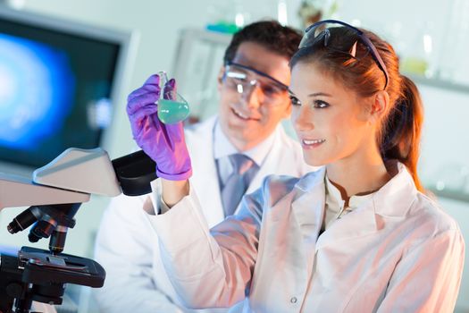 Chemical laboratory scene: attractive young student and her post doctoral supervisor scientist observing the green indikator solution color shift in glass flask.