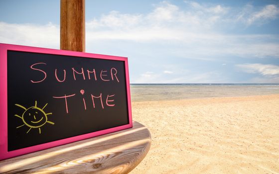 In the picture a beach umbrella which is small blackboard with the words " Summer Time " in a beach in the afternoon .
