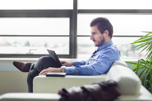 Portrait of successful young businessman in bright modern office focused on work on his laptop computer wearing headphones. Side view. Business and entrepreneurship concept.