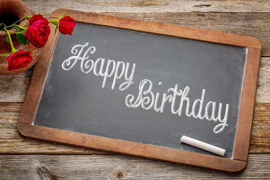 Happy Birthday greetings  - white chalk text on a vintage slate blackboard with red roses against rustic wood