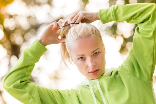 Portrait of attractive confident sporty woman wearing fashionable green sportswear  making her hair in ponytail before training in nature. Active sporty lifestyle concept.