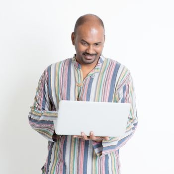 Portrait of mature casual business Indian male using laptop computer, standing on plain background with shadow.