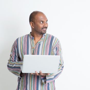 Portrait of mature casual business Indian man using laptop computer, looking at side copy space, standing on plain background with shadow.