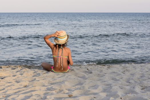 Little girl on the beach at sunset, with har hands blocking a large hat.