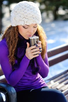 Beautiful girl in white hat sitting on a bench with a cup of coffee in the winter