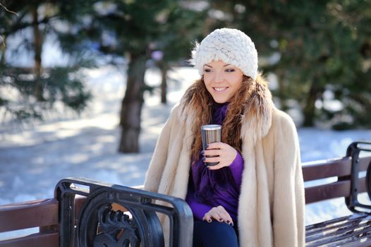 brunette girl in white hat sitting on a bench with a cup in the winter