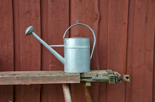 Metal watering can on a bench besides red wooden farm house wall