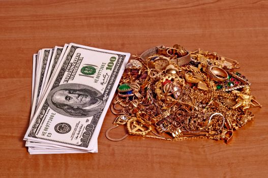 Horizontal shot of a stack of hundred dollar bills next to a pile of gold jewelry on a wood counter top.