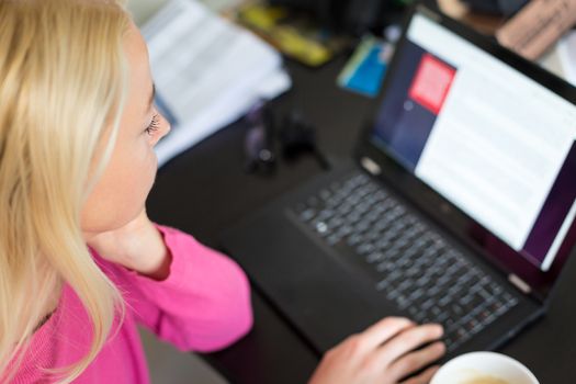 Female freelancer in her casual home clothing remotly working on laptop computer from her home while drinking her morning cup coffee. View from above.