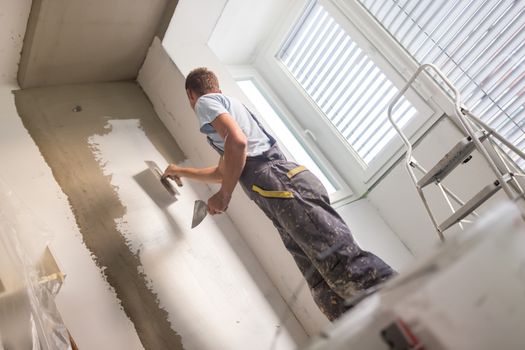 Thirty years old manual worker with wall plastering tools inside a house. Plasterer renovating indoor walls and ceilings with float and plaster.