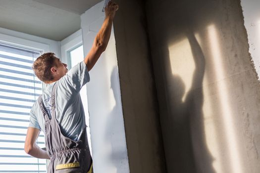 Thirty years old manual worker with wall plastering tools inside a house. Plasterer renovating indoor walls and ceilings with float and plaster.