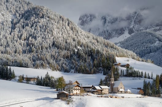 The small village of St. Magdalena or Santa Maddalena with its church covered in snow and with the Dolomites mountains covered in the clouds in the Val di Funes Valley (Villnoesstal) in South Tyrol in Italy in winter.