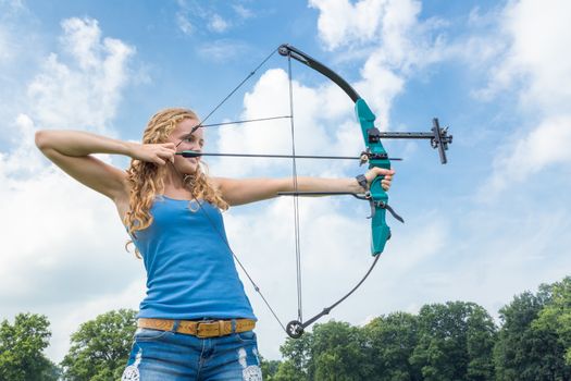 Blonde caucasian girl shooting with arrow and compound bow outdoors on sunny day