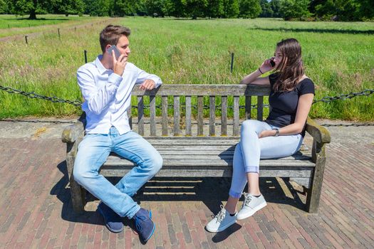 Young attractive caucasian couple on bench calling each other with  mobile phones
