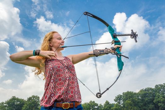 Young woman shooting archery with compound bow against blue sky and white clouds