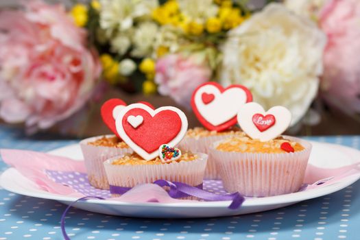 white plate with valentine's day muffins with red and white hearts on a blue with dots (polka dot) table and flowers on the background