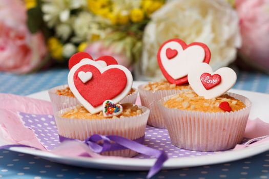 white plate with valentine's day muffins with red and white hearts on a blue with dots (polka dot) table and flowers on the background