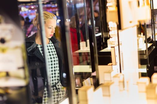 Beautiful blonde lady standing in front of showcase in beauty store, admiring new perfume collection.